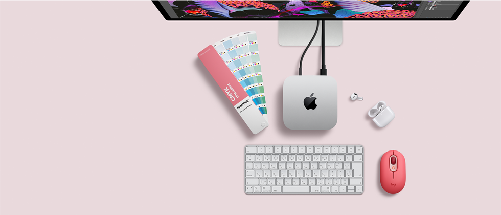 Mac miniを上から見た図。シルバーのアルミニウム製、丸みを帯びた四隅、中央に光沢のあるブラックのAppleのロゴ。コードで接続されている。上部にStudio Displayの一部分が見えている。ケースが開いた状態のAirPods 4、パントンのカラースウォッチ、テンキー付きキーボードとマウス