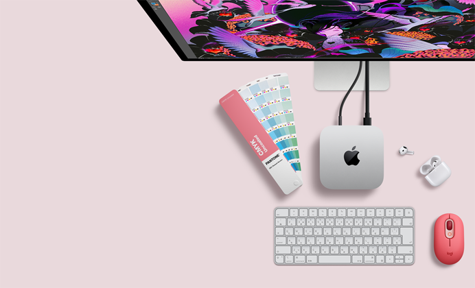 Mac miniを上から見た図。シルバーのアルミニウム製、丸みを帯びた四隅、中央に光沢のあるブラックのAppleのロゴ。コードで接続されている。上部にStudio Displayの一部分が見えている。ケースが開いた状態のAirPods 4、パントンのカラースウォッチ、テンキー付きキーボードとマウス